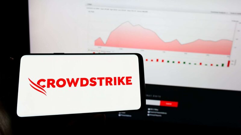 Celular com logotipo da Crowdstrike na tela e, ao fundo, computador com gráfico da empresa aberto