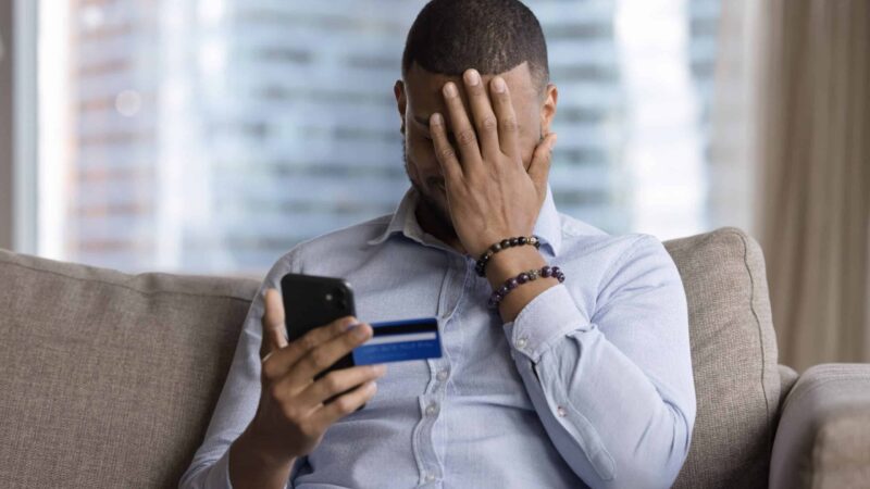 Homem com uma mão no rosto e, na outra, segurando um cartão e um celular